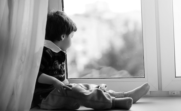 O menino está lendo um livro. A criança se senta à janela e se prepara para as aulas. Menino com um livro nas mãos está sentado no parapeito da janela.