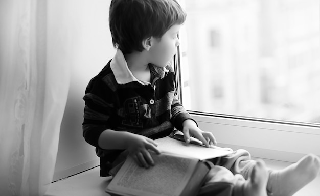 O menino está lendo um livro. A criança se senta à janela e se prepara para as aulas. Menino com um livro nas mãos está sentado no parapeito da janela.