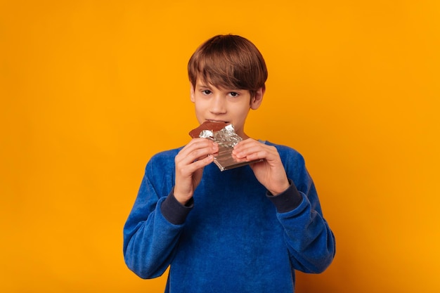 O menino está segurando e mordendo uma barra de chocolate sobre fundo amarelo