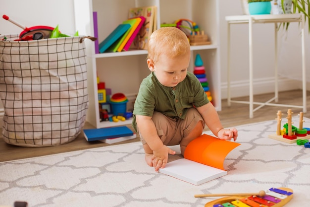 O menino está sentado e olhando para um livro aberto O menino loiro lê um livro no jardim de infância