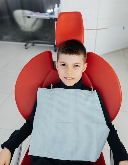 O menino está sentado em uma cadeira odontológica vermelha e sorrindo na odontologia branca moderna Tratamento e prevenção de cárie desde a infância Odontologia moderna e próteses
