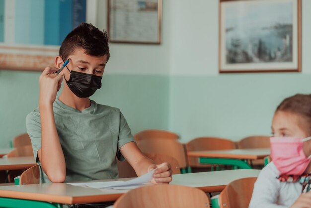 O menino está sentado em uma mesa da escola e usando uma máscara no rosto contra a proteção do vírus corona Nova educação normal durante a pandemia de Covid19 Foco seletivoFoto de alta qualidade