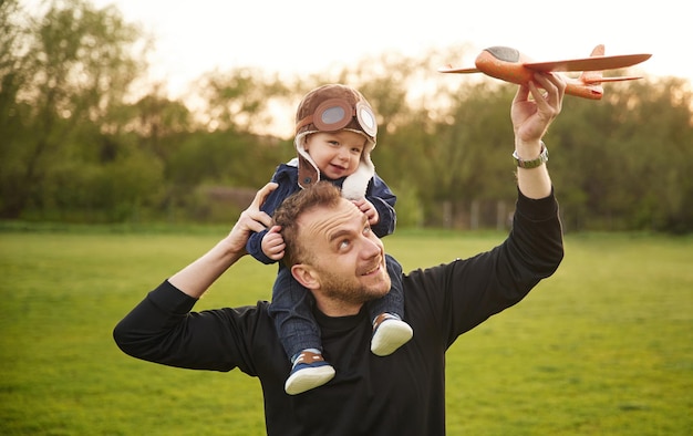 O menino está sentado nos ombros do pai com o seu filhozinho a brincar com um avião de brinquedo no campo.