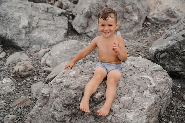 O menino está sentado sorrindo e tomando sol em grandes pedras na praia perto do mar