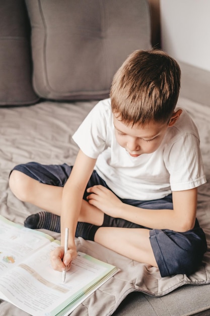 O menino faz a lição de casa sentado no sofá cinza. Um adolescente caucasiano escreve em uma pasta de trabalho no sofá. Ensino doméstico a um menino de 9 anos. Aprendizagem grátis