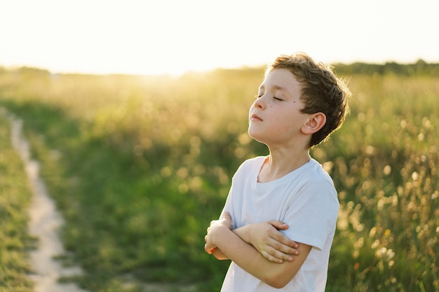 O menino fechou os olhos e rezou em um campo ao pôr do sol mãos dobradas no conceito de oração para a espiritualidade da fé e religião