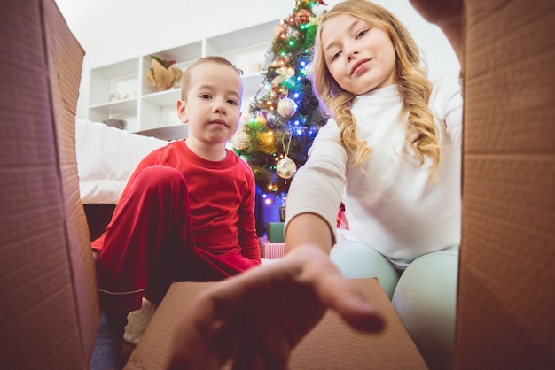 O menino feliz e uma menina abrem a caixa no fundo da árvore de natal