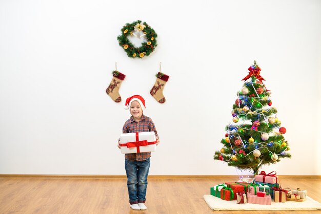 O menino feliz fica perto da árvore de natal e segura uma caixa de presente