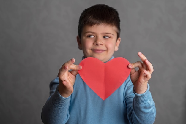 O menino mostra o coração de papel vermelho menino desvia os olhos para o lado criança segura um cartão de dia dos namorados de papel vermelho