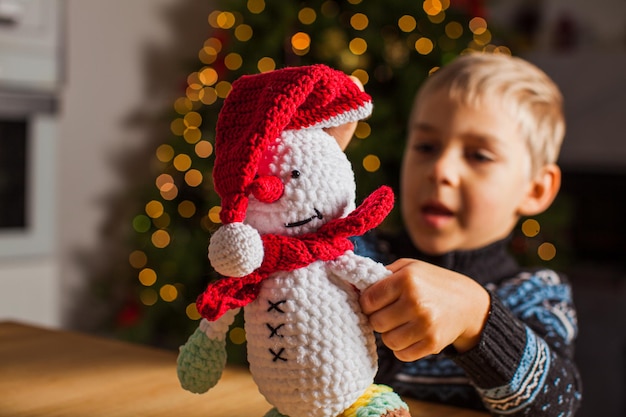 O menino pré-escolar está brincando com um boneco de neve de crochê no contexto da árvore de Natal