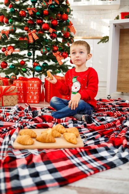 O menino que se senta perto da árvore de Natal para o ano novo. Decoração de Natal com presentes, uma criança perto da árvore de Natal come croissants e sorri