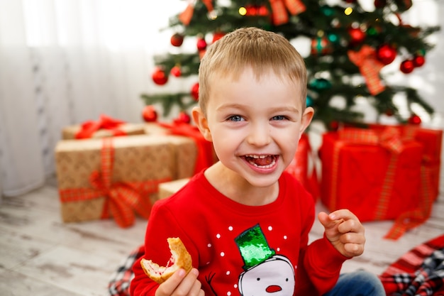 O menino que se senta perto da árvore de Natal para o ano novo. Decoração de Natal com presentes, uma criança perto da árvore de Natal come croissants e sorri