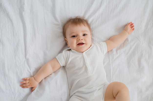 O menino saudável da criança sorri na roupa branca encontra-se na cama com linho branco