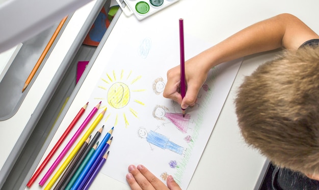 O menino se senta à mesa e desenha com lápis de cor