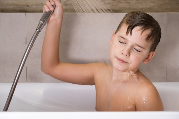 O menino segurando o chuveiro e lavando a cabeça no banheiro. infância saudável e feliz