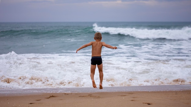 O menino senta-se na costa do oceano com os braços abertos para o vento e as ondas Tempestade na coragem do verão