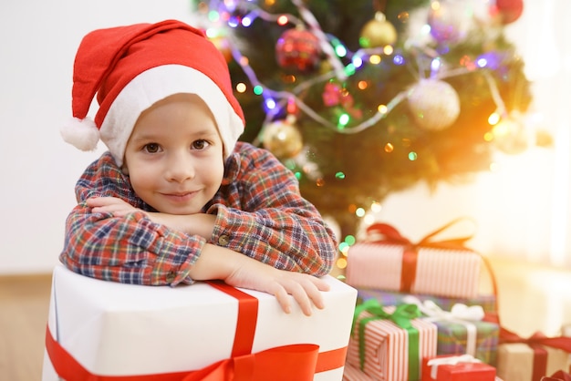 O menino sorridente sentado com uma caixa de presente perto da árvore de Natal em um fundo ensolarado