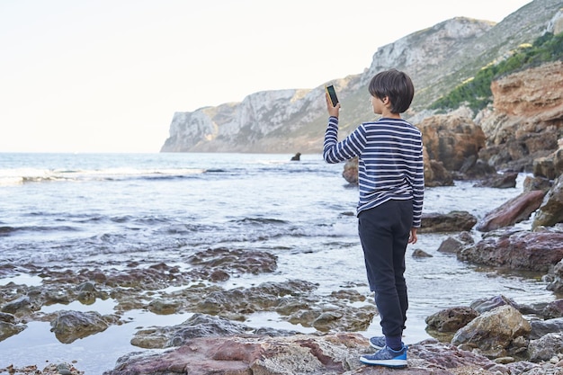 O menino tirando uma foto com seu celular em uma pedra à beira-mar