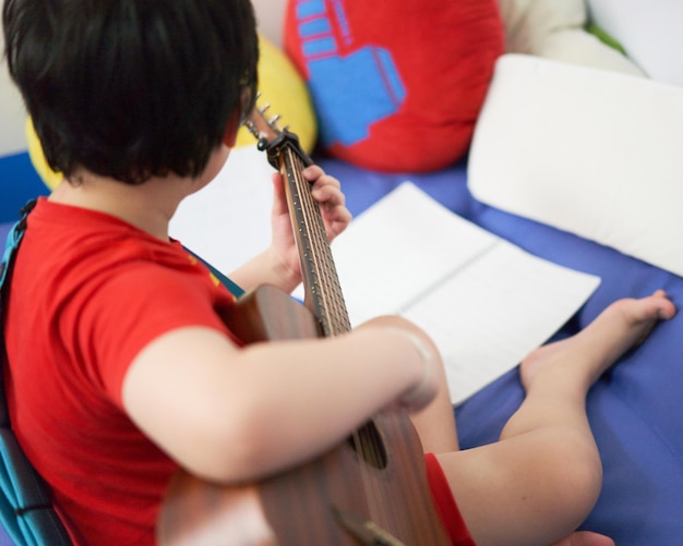 O menino toca violão com a mão esquerda, uma música sozinho, no sofá, no quarto, lendo a partitura.