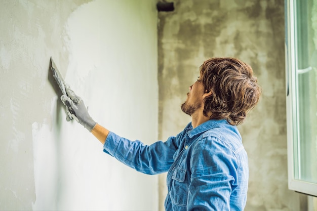 O mestre está aplicando massa branca em uma parede e manchando com espátula em uma sala de renovação de casa durante o dia