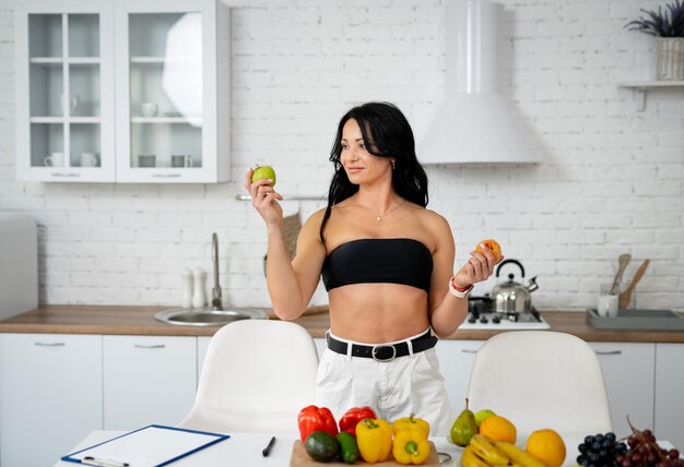 Esta o esta mujer caucásica morena bonita parada cerca de la mesa con comida saludable y tomando decisiones mientras mira la manzana verde Concepto de comida de dieta saludable