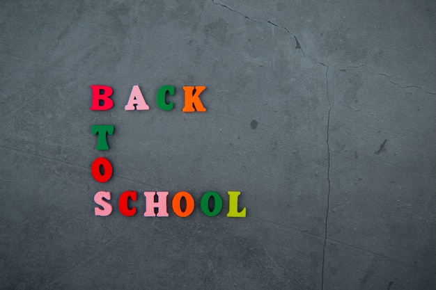 O multicolorido de volta à palavra escola é feito de letras de madeira em uma parede rebocada cinza.