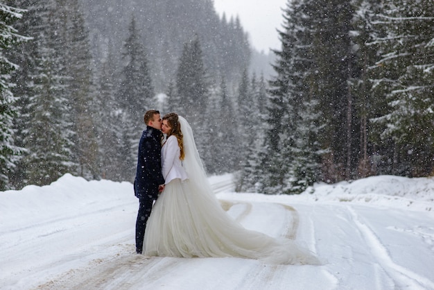 O noivo beija a noiva no contexto de uma floresta de abetos nevados. nevando. casamento de inverno.