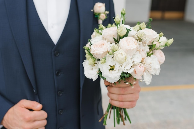 O noivo de terno azul segura na mão um delicado buquê de casamento para sua amada noiva