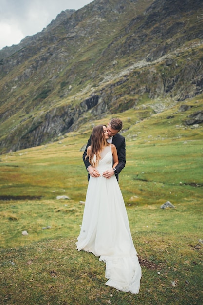 O noivo de terno e a noiva em um lindo branco posando contra a paisagem verde da montanha cas...