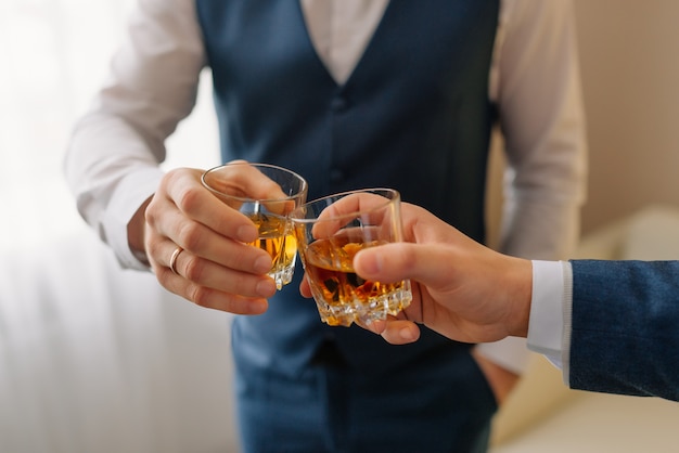 O noivo e o padrinho bebendo um uísque, comemorando o dia do casamento. Mãos de homem segurando copos com álcool e brindar. grupo de homens torcendo e tilintando com bebidas na recepção do casamento