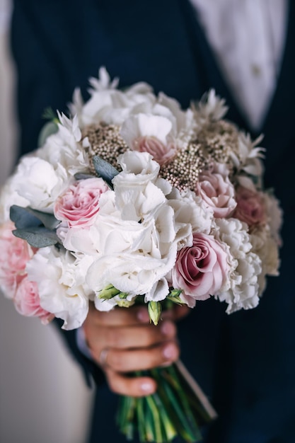 O noivo em uma jaqueta escura segura um buquê de uma noiva de flores rosa e brancas