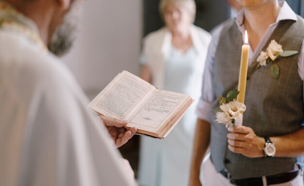 O padre com a bíblia fica ao lado do noivo com uma vela na mão