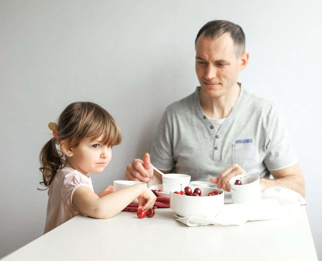O pai alimenta uma filhinha bonitinha com um café da manhã saudável em casa em um fundo claro. Família feliz