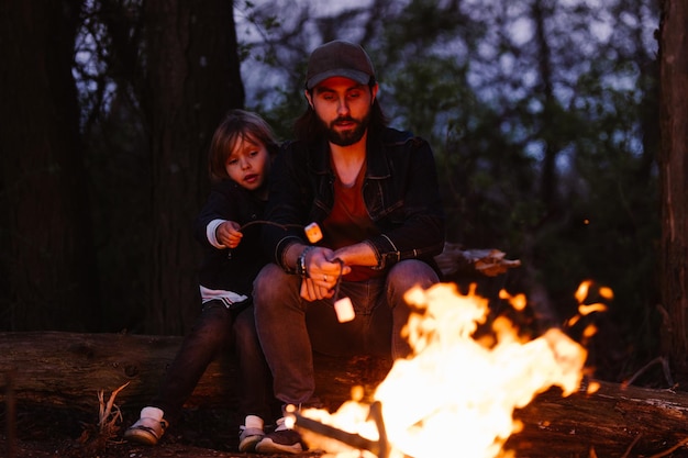 O pai e o filho sentados nos troncos da floresta e assando marshmallows no fogo