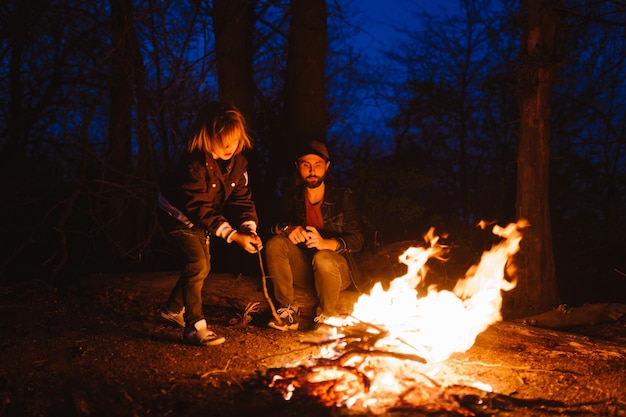 O pai e o filho sentados nos troncos da floresta e assando marshmallows no fogo