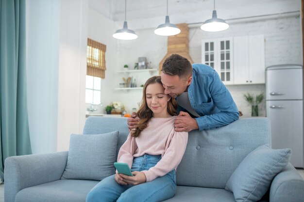 O pai está de pé atrás do sofá, abraçando os ombros de uma filha que está sentada no sofá, ambas olhando para a tela do smartphone.