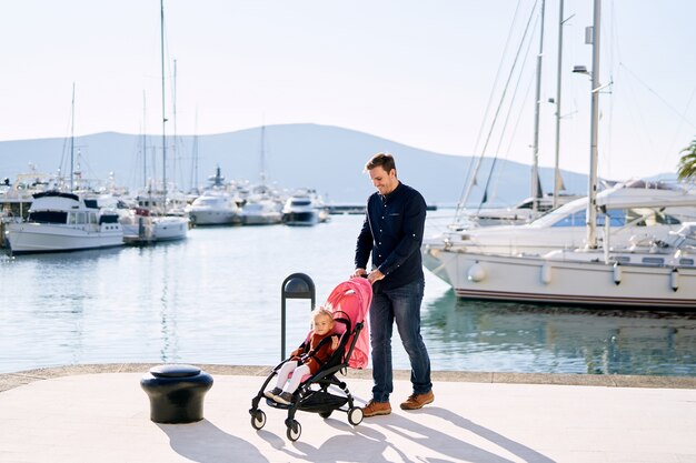 O pai está empurrando um carrinho rosa com sua filha sentada dentro de uma marina de barcos.