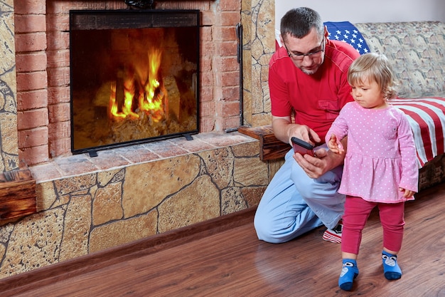 O pai está mostrando seu smartphone para a filha, eles estão perto da lareira e a garota está interessada no dispositivo.