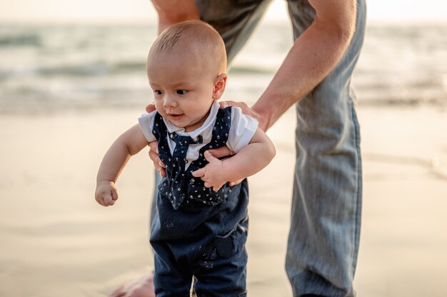 O pai estava segurando seu filho para aprender a andar na praia ao pôr do sol. Família de férias na praia no verão, viagens, férias e conceito de estilo de vida