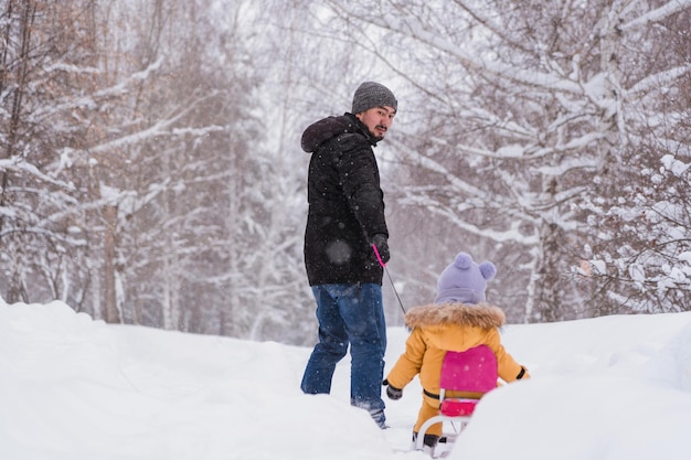 O pai puxa um gato de neve infantil com uma criança sentada nele por 1217 meses através do parque de inverno durante um foco seletivo de queda de neve Fundos desfocados