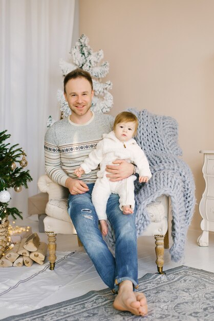 O pai segura a filha nos braços e se senta em uma cadeira perto da árvore de Natal