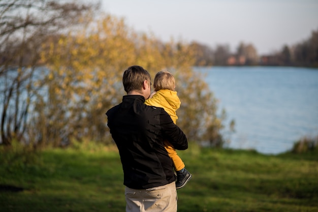 O pai segura o filho com uma jaqueta amarela nos braços e olha para o lago