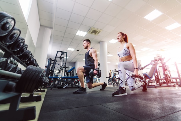 O par saudável no esporte veste levantar pesos no gym. Mulher atraente e homem bonito a fazer exercícios com halteres em pé em pose especial no clube desportivo.