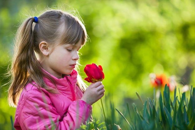 O perfil da menina de sorriso bonita bonito da criança com olhos cinzentos e cabelo longo que cheira a flor vermelha brilhante da tulipa no fundo ensolarado borrado do bokeh do verde do verão.