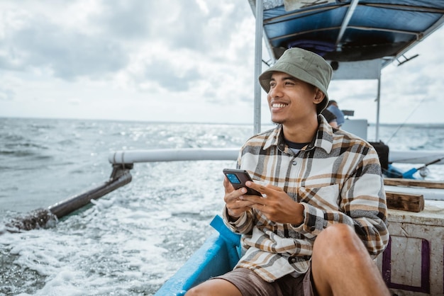 O pescador asiático senta-se relaxado ao usar o smartphone no barco de pesca