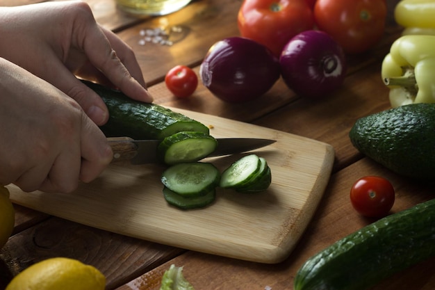 O processo de fatiar vegetais para salada vegetariana. As mãos femininas cortam tomates, pepinos, cebolas, pimentão em uma mesa de madeira.