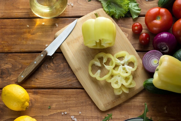 O processo de fatiar vegetais para salada vegetariana. As mãos femininas cortam tomates, pepinos, cebolas, pimentão em uma mesa de madeira.