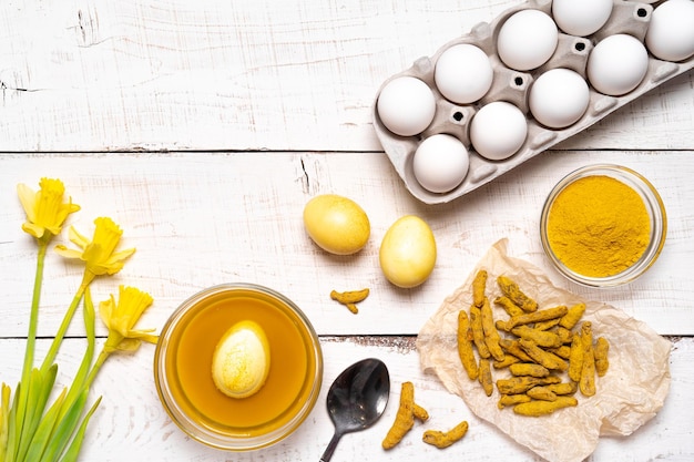 O processo de pintura de ovos de páscoa com corantes vegetais naturais açafrão em uma vista superior de fundo branco de madeira de ovos de galinha e narcisos amarelos