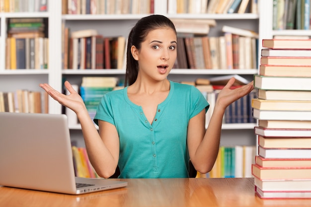 O que escolher? Mulher jovem confusa sentada na mesa da biblioteca com uma pilha de livros e um laptop