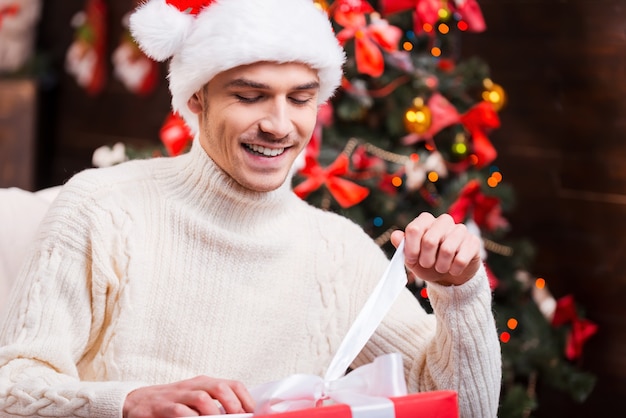 O que esta dentro? Jovem bonito com chapéu de Papai Noel abrindo uma caixa de presente e sorrindo com uma árvore de Natal ao fundo